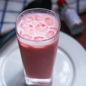 Rose Milk - Rose Milk Syrup A Tall Glass Of Pink, Cold Drink With Ice, Placed On A White Plate, In Dm Traders Organic Store.